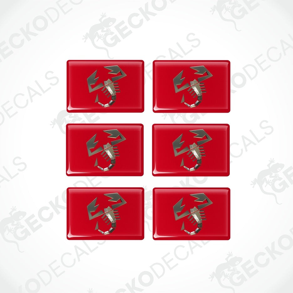 Six glossy red rectangular decals arranged in a 3-by-2 grid on a light gray background. Each decal features a stylized metallic silver and gray emblem resembling a winged gear or mechanical symbol.