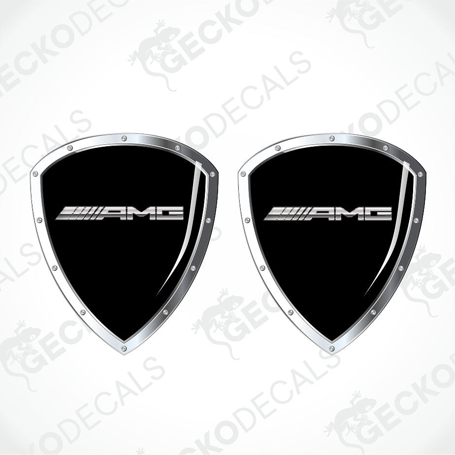 Two black AMG shield-shaped decals with silver borders and the white AMG logo, shown on a light gray background with faint watermark text.