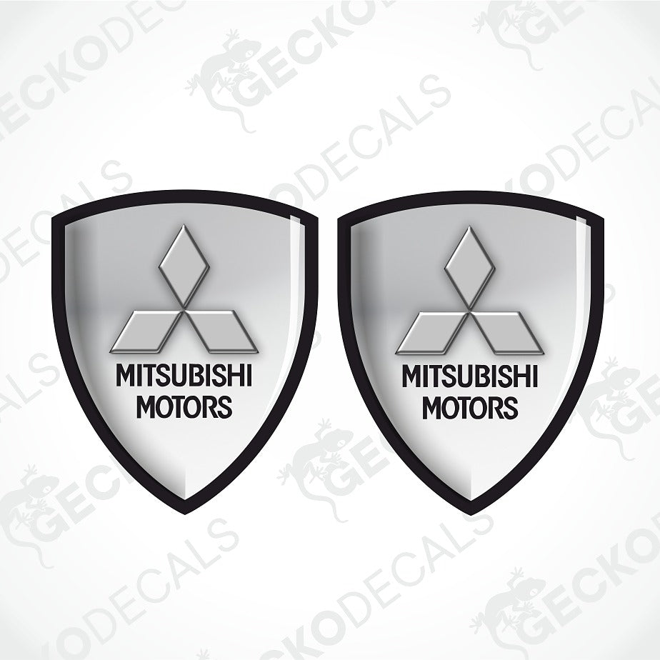 Two identical Mitsubishi Motors shield-shaped decals on a light gray background with diagonal watermarks. Each shield is silver-gray with a black border and the Mitsubishi logo and text.