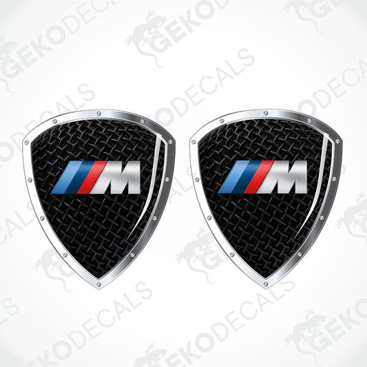 Two identical BMW M shield-shaped decals on a light gray background with diagonal watermark text. Each decal has a black diamond-textured center, a silver border, and the blue, red, and silver BMW M logo.