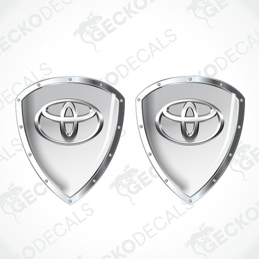 Two identical silver shield-shaped decals featuring the Toyota logo, displayed side-by-side on a light gray background with diagonal watermarks.