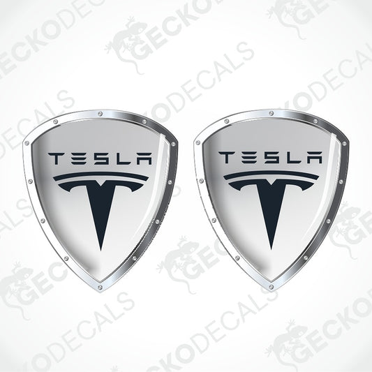 Two identical Tesla shield-shaped decals on a light gray background with diagonal watermark text. Each decal has a silver metallic border, a white center, the TESLA wordmark, and the Tesla T logo in black.