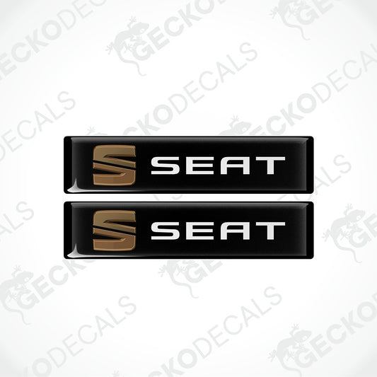 Two glossy black rectangular Seat decal stickers with a metallic gold 'S' logo and white 'SEAT' text, shown on a white background with diagonal watermarks.