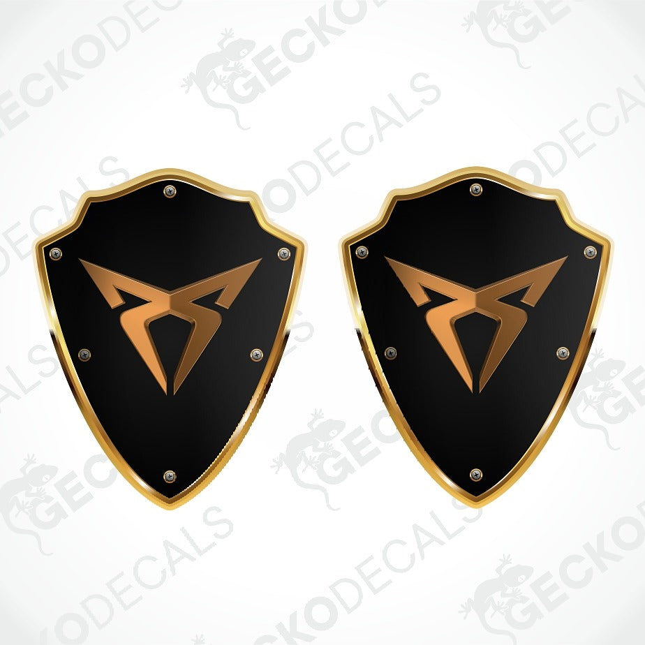 Two identical black shield-shaped decals with gold borders and a gold stylized logo in the center, shown on a white background with faint diagonal watermark text.
