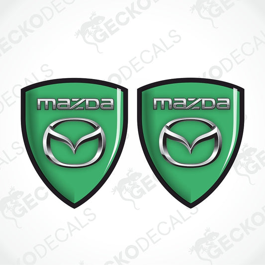 Two green Mazda shield-shaped decals with black borders and silver Mazda logos on a white background with faint watermark text.