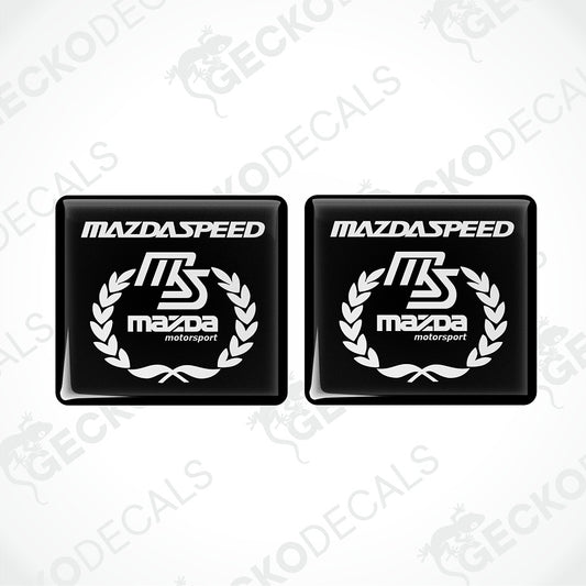 Two black square decals with rounded corners featuring the white Mazda Speed and Mazda motorsport logo, shown on a light gray background with diagonal watermarks.