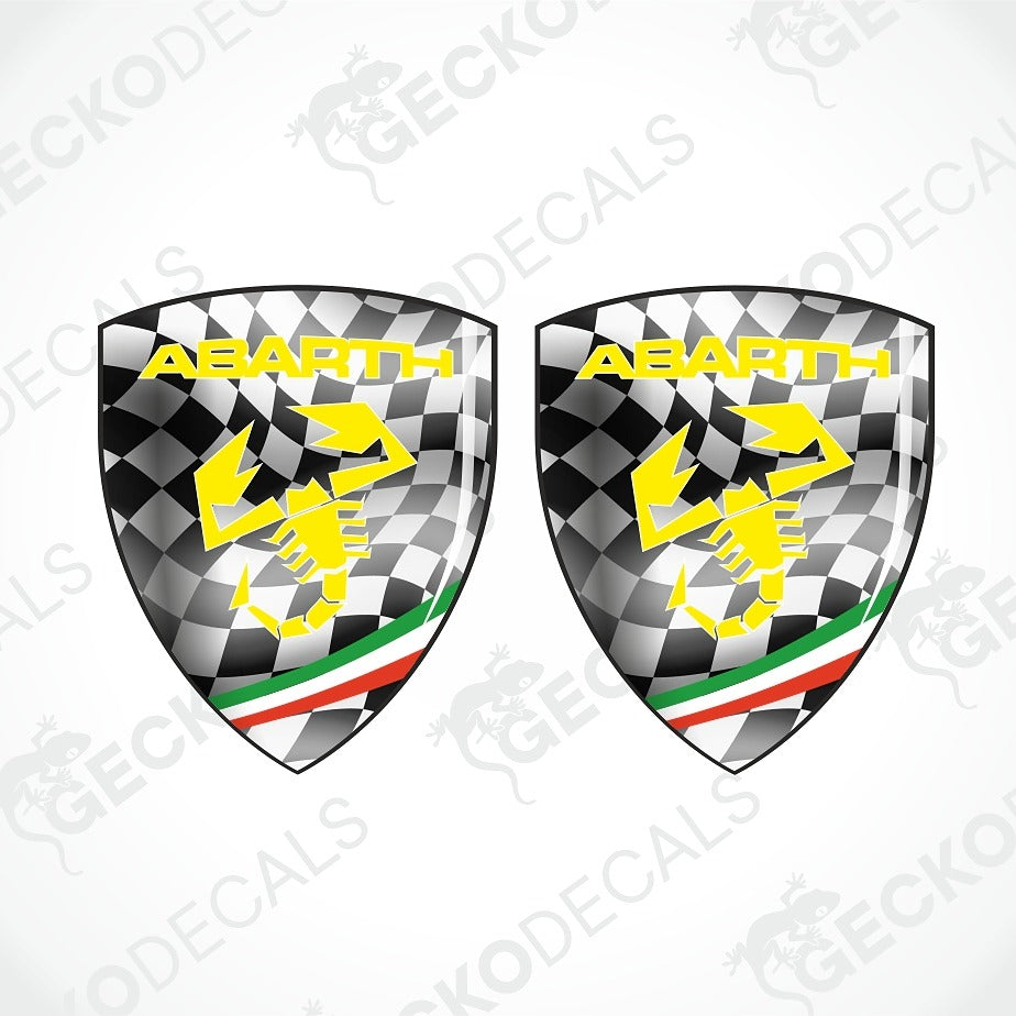 Two Abarth shield-shaped decals with a black-and-white checkered pattern, yellow Abarth text and scorpion graphic, and green, white, and red stripes near the bottom, shown on a light gray background with faint watermark text.