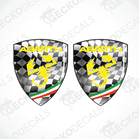 Two Abarth shield-shaped decals with a black-and-white checkered pattern, yellow Abarth text and scorpion graphic, and green, white, and red stripes near the bottom, shown on a light gray background with faint watermark text.