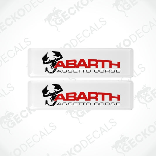 Two rectangular Abarth Assetto Corse decals stacked vertically on a light gray background. The decals are white with a black wolf logo and red and black text.