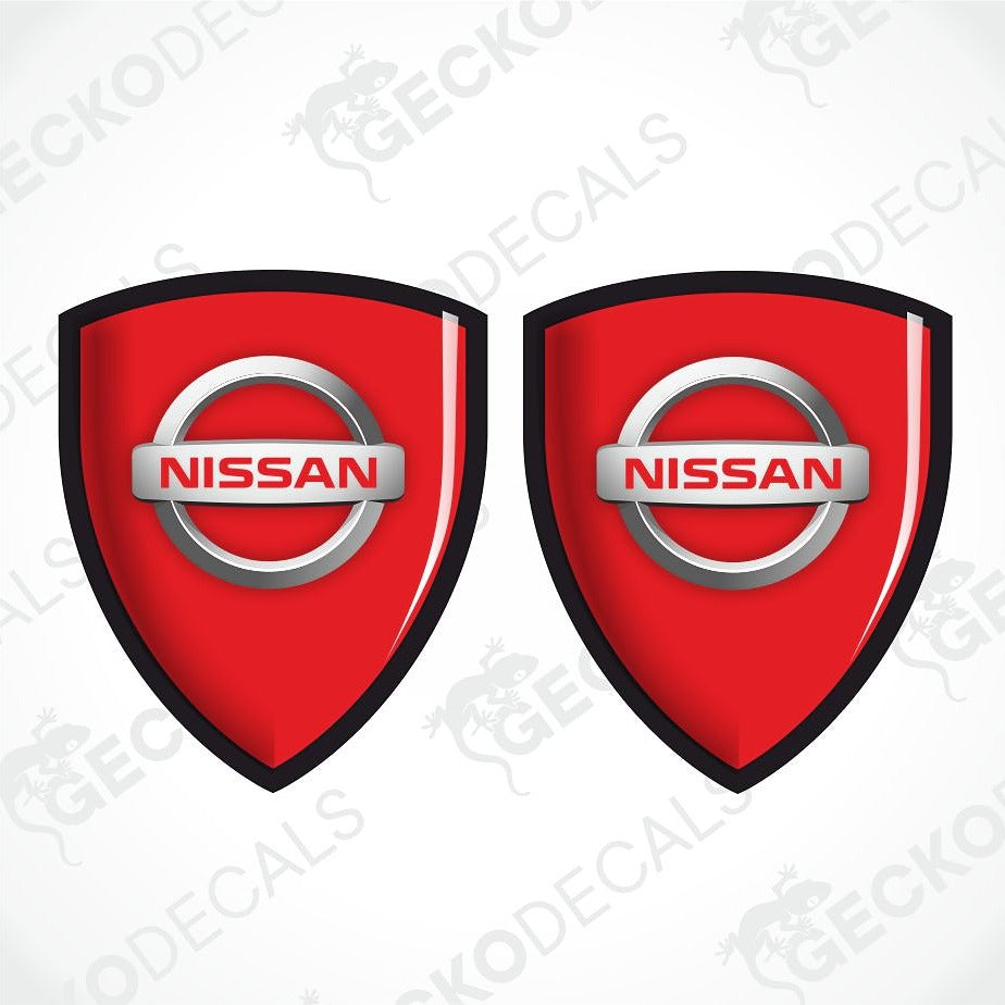 Two red Nissan shield-shaped decals with black borders and silver Nissan logos, shown on a white background with a faint watermark pattern.