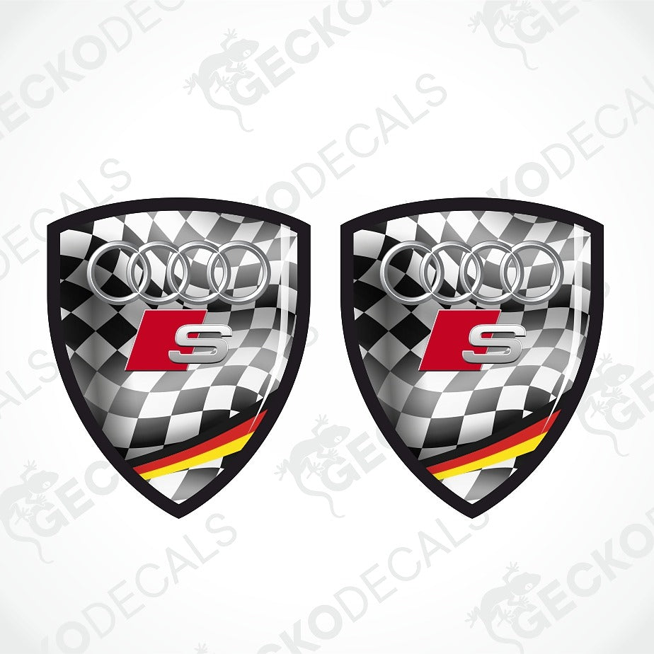 Two identical shield-shaped Audi S decal designs on a light gray background with diagonal watermark text. The decal features a black and white checkered flag pattern, the silver Audi four rings, a red and silver S logo, and a black, red, and yellow German flag stripe at the bottom.
