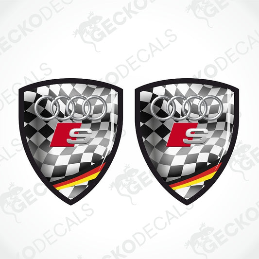 Two identical shield-shaped Audi S decal designs on a light gray background with diagonal watermark text. The decal features a black and white checkered flag pattern, the silver Audi four rings, a red and silver S logo, and a black, red, and yellow German flag stripe at the bottom.