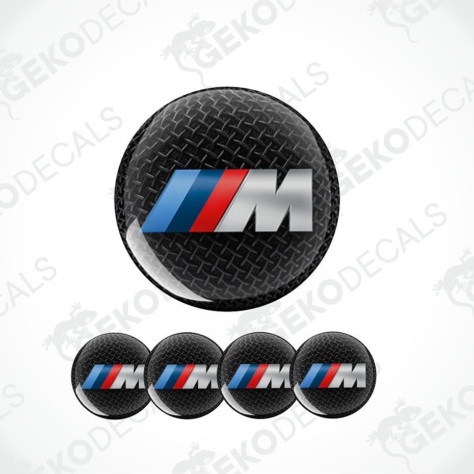 Four round black carbon-fiber-textured BMW M logo decals on a white background, with one larger decal above and four smaller matching decals below. The decal features the blue, red, and silver BMW M tri-color stripe design over a black diamond-plate style texture.