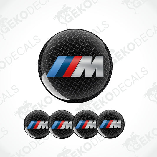 Four round black carbon-fiber-textured BMW M logo decals on a white background, with one larger decal above and four smaller matching decals below. The decal features the blue, red, and silver BMW M tri-color stripe design over a black diamond-plate style texture.