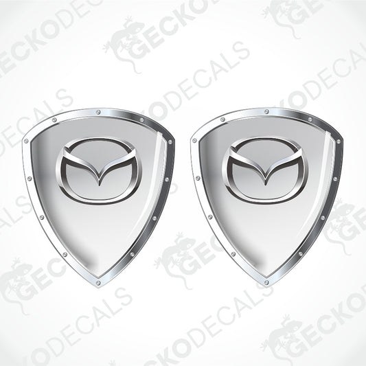 Two identical metallic silver shield-shaped decals with the Mazda logo in the center, shown on a light gray background with faint GECKODECALS watermarks.