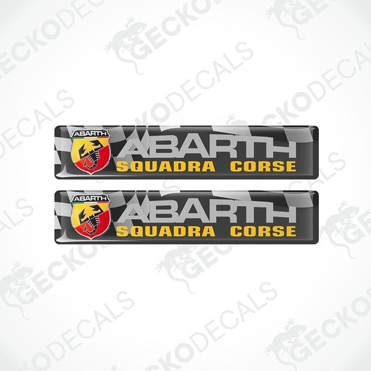 Two Abarth Squadra Corse decal stickers shown on a light gray background with diagonal watermark text. The decals feature a black background with a gray checkered pattern, large silver-gray 'ABARTH' text, yellow 'SQUADRA CORSE' text, and the red and yellow Abarth logo.
