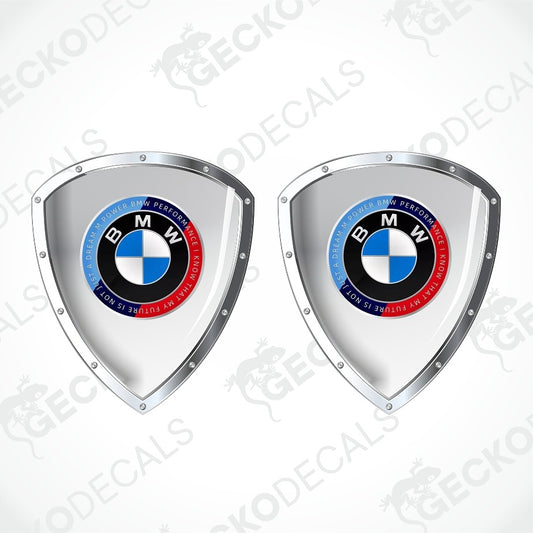 Two silver shield-shaped decals featuring the BMW roundel logo, shown side by side on a light gray background with diagonal watermark text.