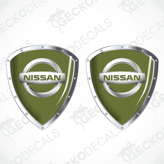 Two identical Nissan shield-shaped decals on a white background with diagonal watermark text. The decals feature an olive green center with a silver metallic border and a silver horizontal bar displaying the NISSAN logo.