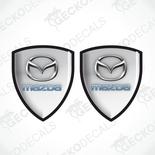 Two identical silver Mazda shield-shaped decals with black borders and blue 'mazda' text, shown on a light gray background with repeated GECKODECAL watermarks.