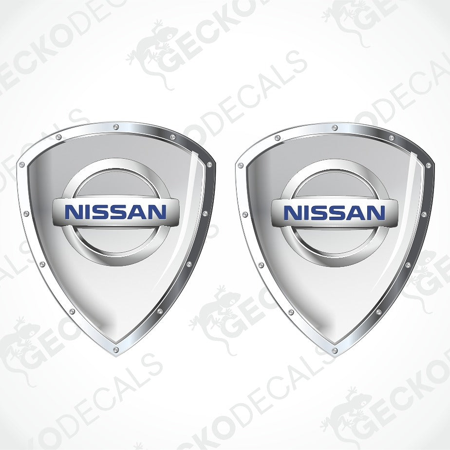Two Nissan shield-shaped decal stickers with metallic silver borders and the blue Nissan logo in the center, shown on a light gray background with a faint watermark pattern.