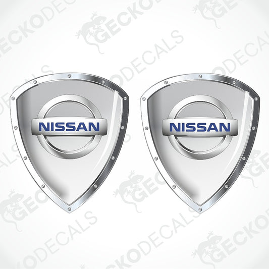 Two Nissan shield-shaped decal stickers with metallic silver borders and the blue Nissan logo in the center, shown on a light gray background with a faint watermark pattern.