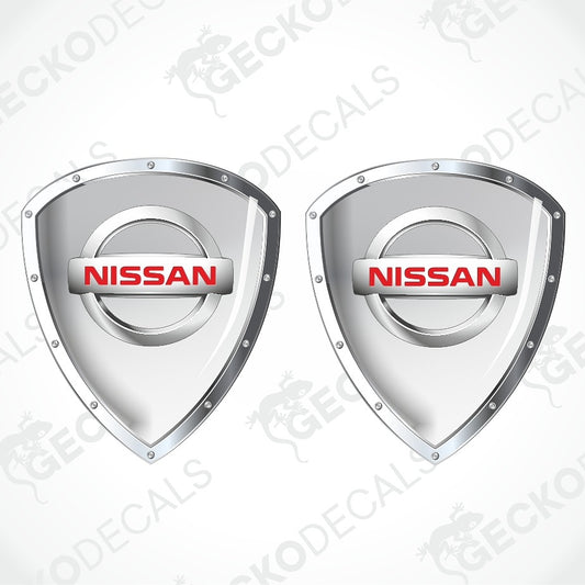 Two silver Nissan shield-shaped decal stickers with red NISSAN logos, shown on a light gray background with diagonal watermarks.