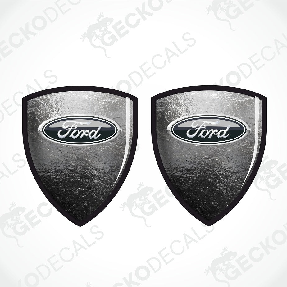 Two Ford shield-shaped decals with a textured metallic silver and black finish, featuring the classic oval Ford logo in the center, shown on a light gray background with a faint watermark pattern.
