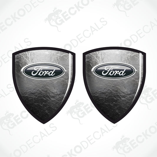 Two Ford shield-shaped decals with a textured metallic silver and black finish, featuring the classic oval Ford logo in the center, shown on a light gray background with a faint watermark pattern.