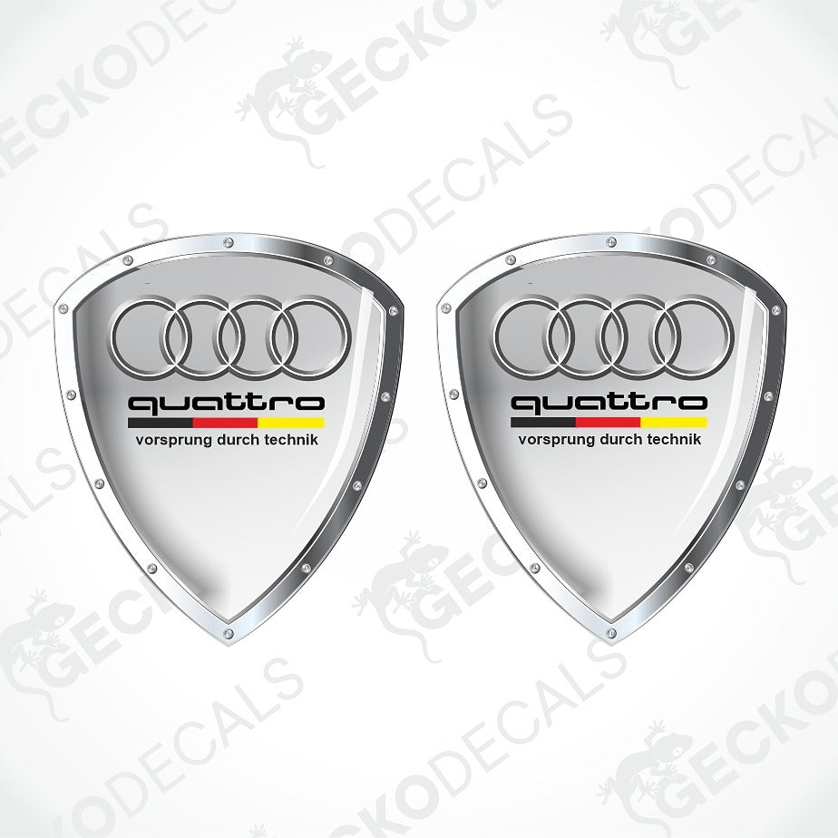 Two silver Audi quattro shield-shaped decals with the four-ring logo and black 'quattro' text on a white background with faint watermark overlays.
