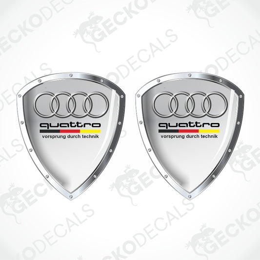 Two silver Audi quattro shield-shaped decals with the four-ring logo and black 'quattro' text on a white background with faint watermark overlays.