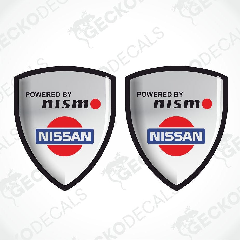 Two identical shield-shaped decals featuring the 'Powered by Nismo' and 'Nissan' logo, displayed side-by-side on a light gray background with diagonal watermarks.