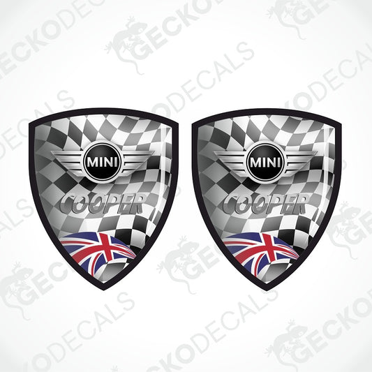 Two identical Mini Cooper shield-shaped decals on a light gray background. Each decal features a black and white checkered flag pattern, a circular black Mini logo with silver wings, the word 'COOPER' in stylized lettering, and a Union Jack flag at the bottom.