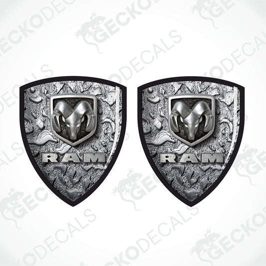 Two matching Ram shield-shaped decals shown side by side on a light gray background. Each decal features a silver ram head logo above the word RAM in raised lettering, with an ornate textured gray background and black border.