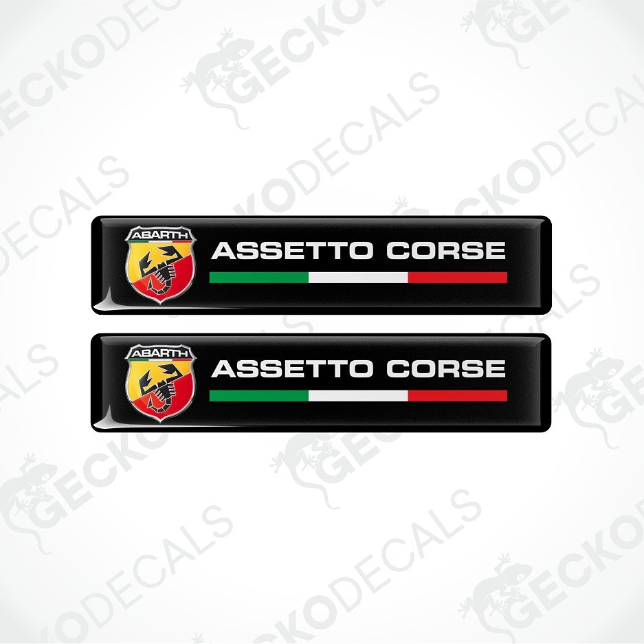 Two black rectangular Abarth Assetto Corse decals with the Abarth logo, white text, and green, white, and red stripes, shown on a light gray background with faint Geekodec watermarks.