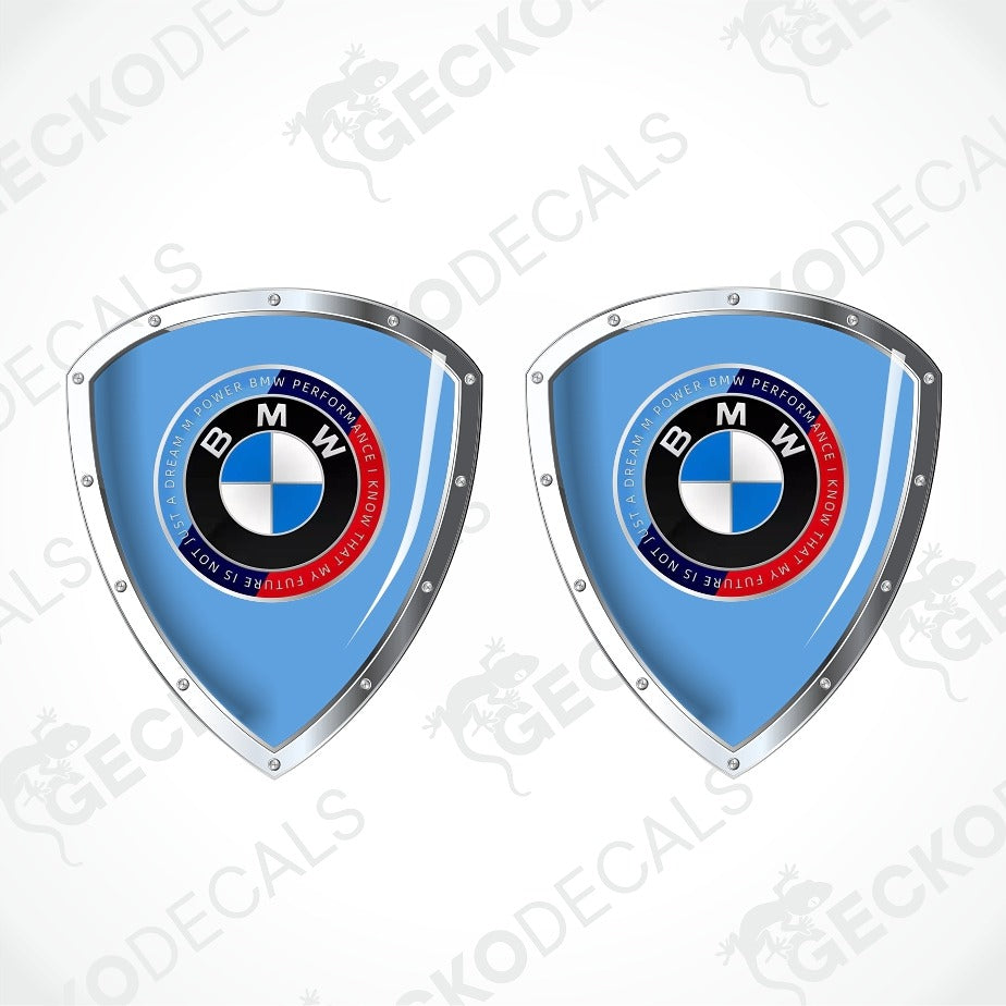 Two identical BMW shield-shaped decals on a light gray background with diagonal watermarks. Each decal has a light blue center, a silver metallic border, and the circular BMW logo in the middle.