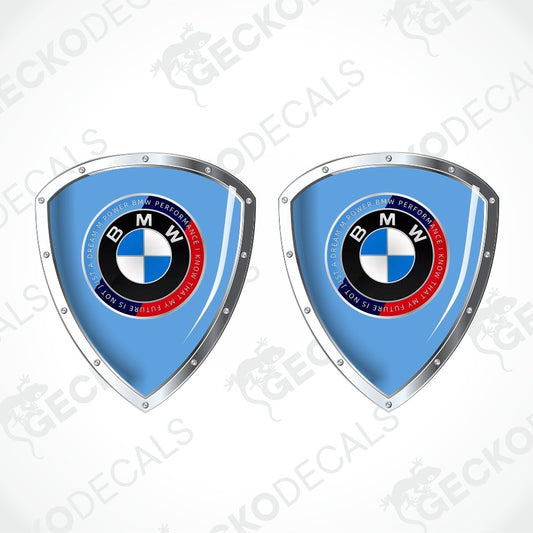 Two identical BMW shield-shaped decals on a light gray background with diagonal watermarks. Each decal has a light blue center, a silver metallic border, and the circular BMW logo in the middle.