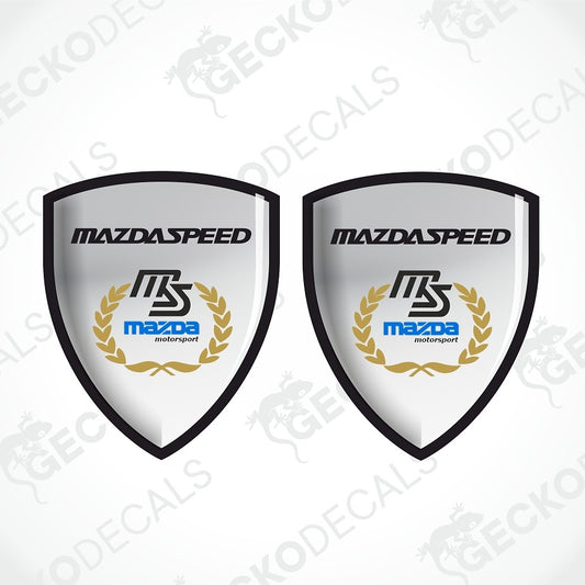 Two identical shield-shaped MazdaSpeed decal designs on a light gray background. Each shield has a black border, a white center, the text 'MAZDASPEED' at the top, a stylized 'MS' logo, the Mazda motorsport wordmark in blue, and a gold laurel wreath.