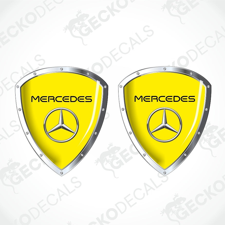 Two Mercedes-Benz shield-shaped decals with bright yellow centers, silver metallic borders, and the Mercedes star logo, shown on a light gray background with faint watermark text.