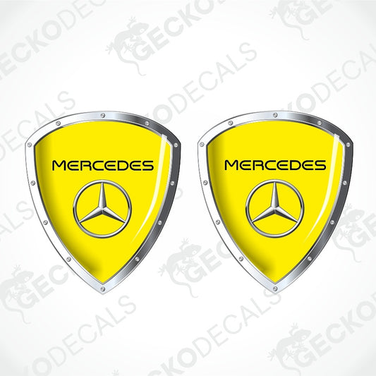 Two Mercedes-Benz shield-shaped decals with bright yellow centers, silver metallic borders, and the Mercedes star logo, shown on a light gray background with faint watermark text.