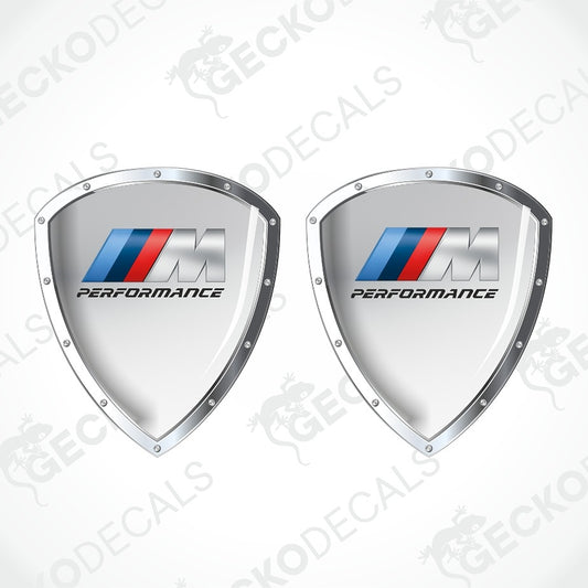 Two identical silver shield-shaped decals with the BMW M Performance logo and text, shown on a white background with diagonal watermark overlays.