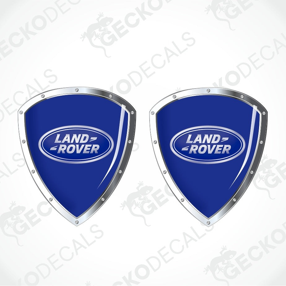 Two blue Land Rover shield-shaped decals with silver metallic borders and the Land Rover logo in the center, shown on a light gray background with faint watermark text.