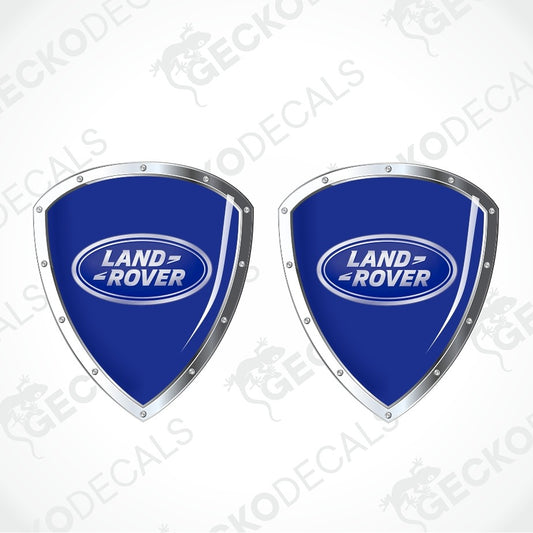 Two blue Land Rover shield-shaped decals with silver metallic borders and the Land Rover logo in the center, shown on a light gray background with faint watermark text.