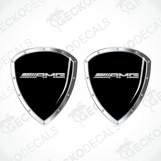 Two black AMG shield-shaped decals with silver borders and the white AMG logo, shown on a light gray background with faint watermark text.