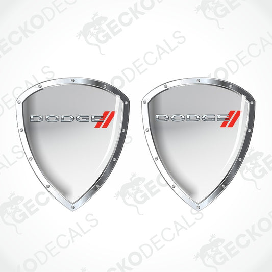 Two silver metallic shield-shaped decal emblems with the DODGE logo and red double stripes, shown on a light gray background with a faint watermark pattern.