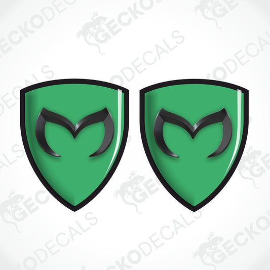 Two identical green shield-shaped decals with black borders and a black stylized 'M' logo in the center, shown on a white background with a faint repeating watermark.