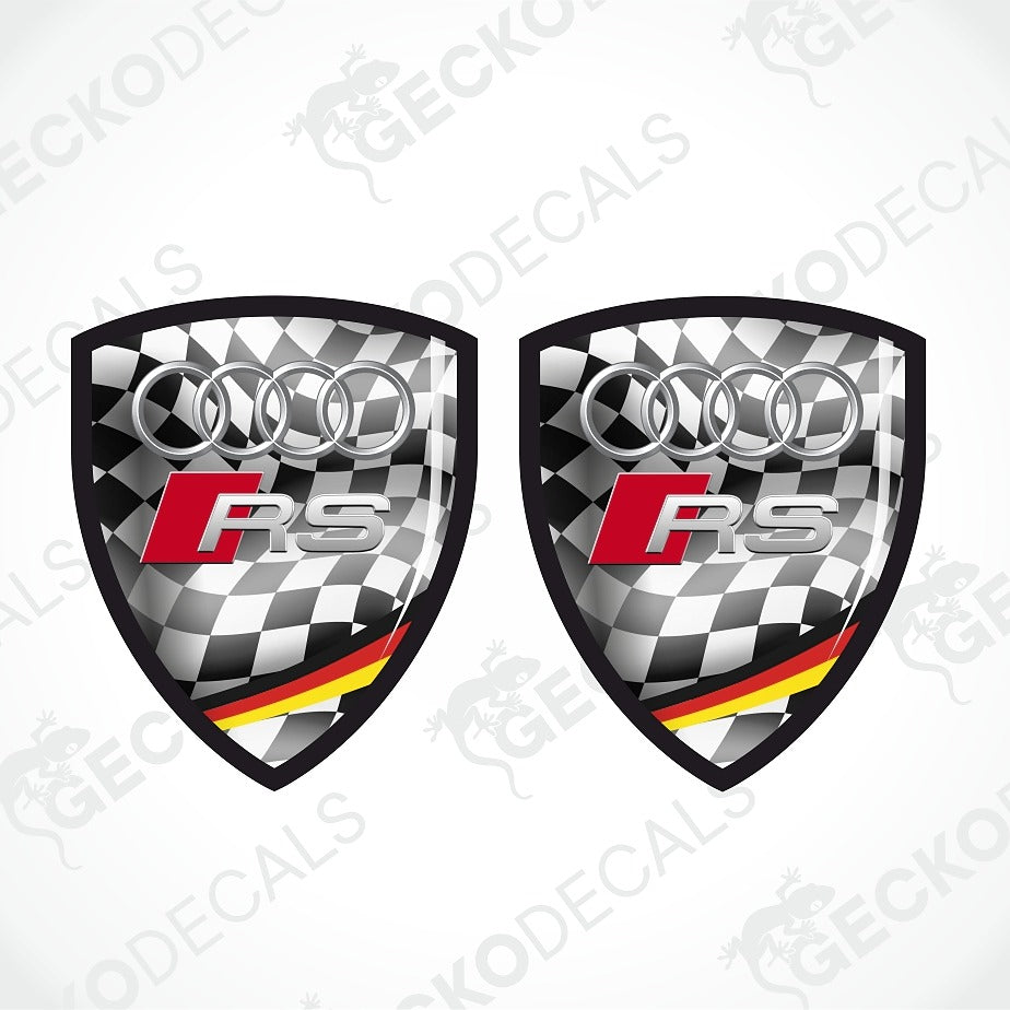 Two identical Audi RS shield-shaped decals on a light gray background with diagonal watermark text. The decals feature a black-and-white checkered pattern, the silver Audi four rings, a red and silver RS logo, and a small German flag stripe in red, black, and yellow at the bottom.