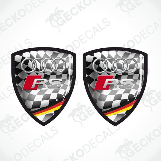 Two identical Audi RS shield-shaped decals on a light gray background with diagonal watermark text. The decals feature a black-and-white checkered pattern, the silver Audi four rings, a red and silver RS logo, and a small German flag stripe in red, black, and yellow at the bottom.