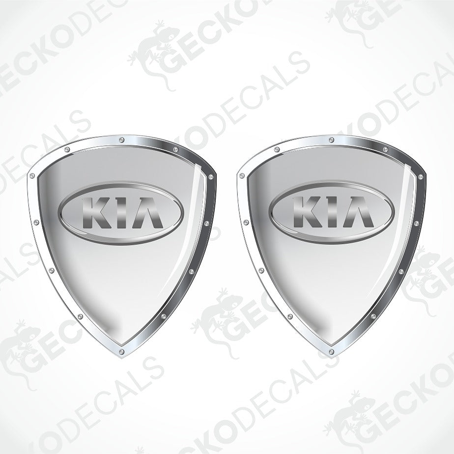 Two metallic silver shield-shaped Kia decal stickers with raised oval Kia logos, shown on a light gray background with diagonal watermark text.