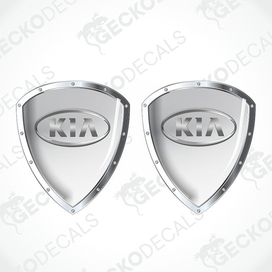 Two metallic silver shield-shaped Kia decal stickers with raised oval Kia logos, shown on a light gray background with diagonal watermark text.