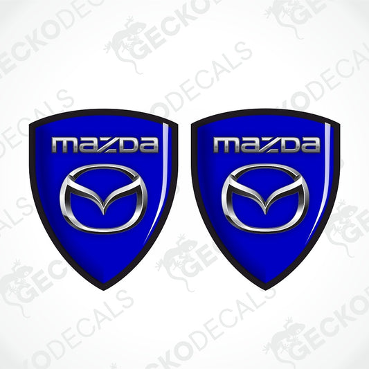 Two identical blue Mazda shield-shaped decals with silver Mazda logo and wordmark, shown on a white background with diagonal watermark text.